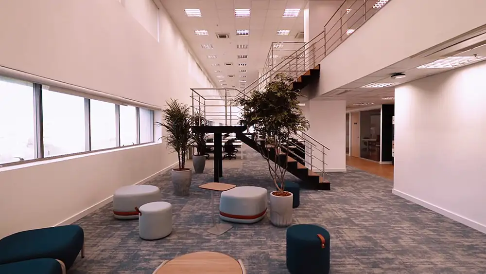 Área colaborativa do escritório da Oceaneering no Rio de Janeiro, projetada pela Custódio. Espaço amplo com escada metálica, mobiliário informal e integração visual, destacando o projeto de arquitetura corporativa e a reforma do escritório.