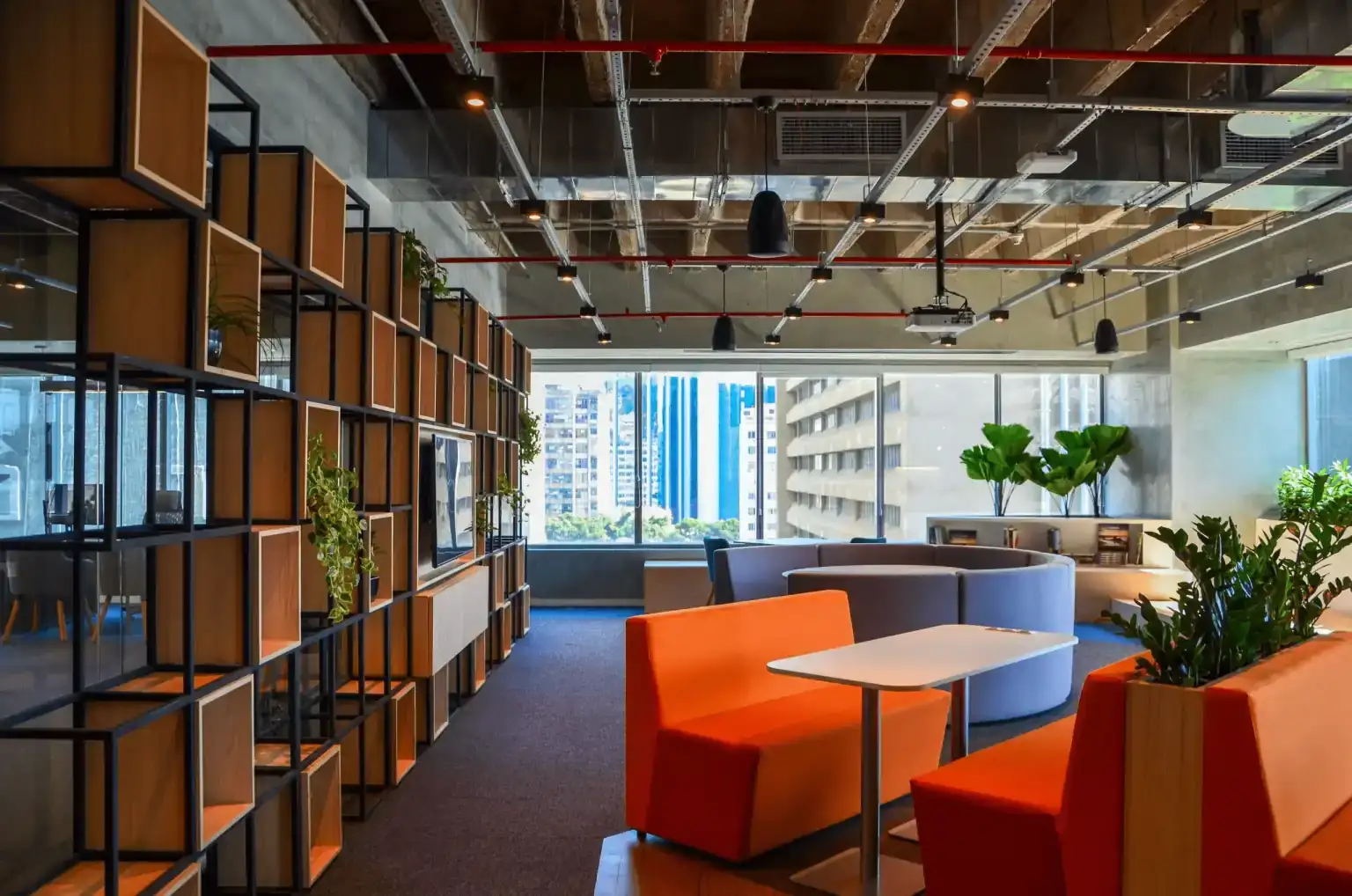 Área de convivência do escritório da EDF projetada pela Custódio, com booths coloridos, estante modular de madeira e metal, iluminação industrial aparente, plantas decorativas e vista urbana através das janelas amplas.