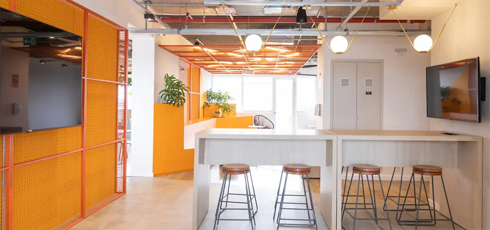 Área de convivência do escritório da Lalamove com mesas altas, bancos industriais e divisórias laranja, destacando o projeto de arquitetura aplicado na obra corporativa e na reforma de escritório com atmosfera moderna.