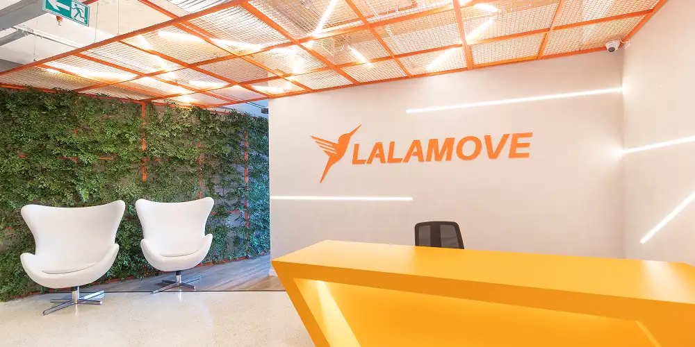 Recepção do escritório da Lalamove com balcão laranja, parede viva e iluminação linear, evidenciando o projeto de arquitetura aplicado na obra corporativa e na reforma de escritório com identidade vibrante.