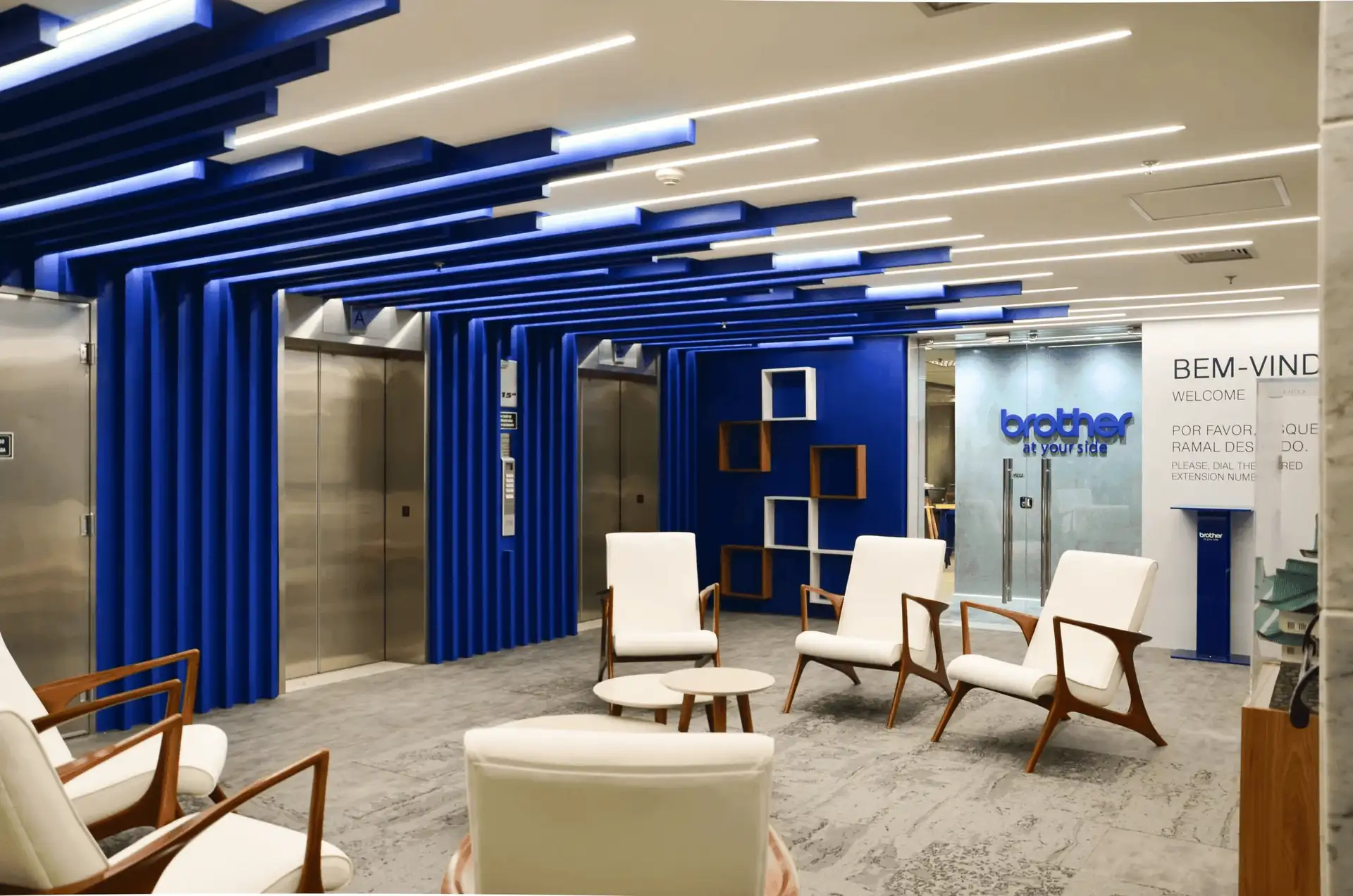 Recepção corporativa da Brother com teto azul geométrico, poltronas brancas e hall de elevadores, destacando projeto de arquitetura moderno, obra corporativa detalhada e reforma de escritório com forte identidade visual.