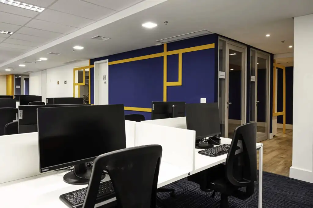 Open space do escritório da Saipem com estações de trabalho brancas, divisórias baixas, salas individuais envidraçadas e parede azul com detalhes amarelos, representando um projeto de arquitetura e reforma corporativa moderna.