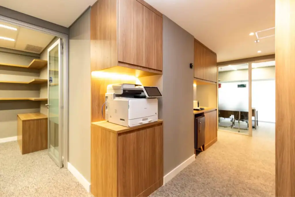 Área de apoio do escritório Villemor Amaral com armários em madeira, copa compacta e impressora embutida, destacando projeto de arquitetura corporativa e reforma de escritório funcional e elegante.