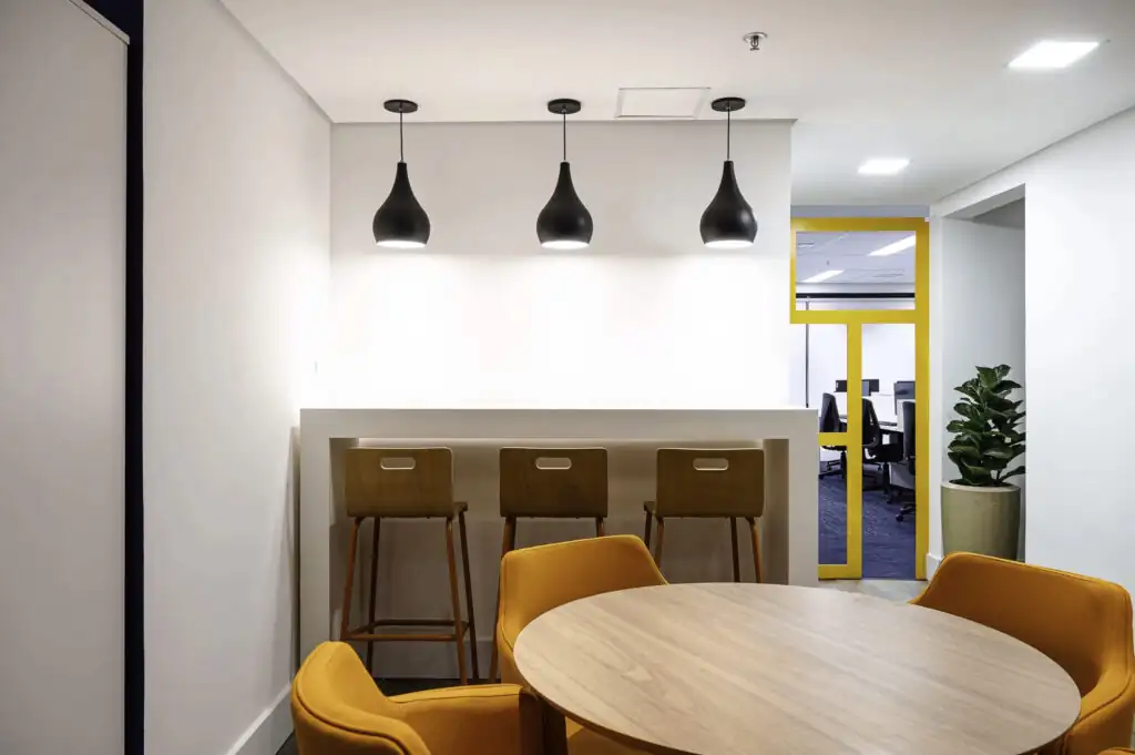 Área de convivência do escritório da Saipem com mesa redonda, cadeiras estofadas, bancada elevada com banquetas e porta amarela ao fundo, destacando projeto de arquitetura e reforma corporativa.