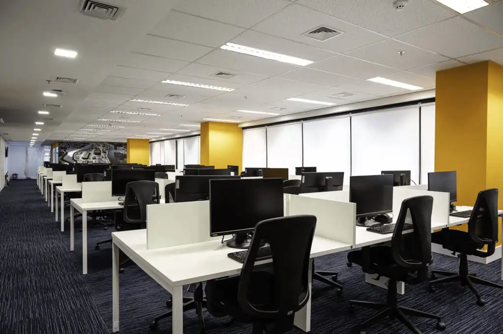 Open space operacional do escritório da Saipem com estações de trabalho alinhadas, divisórias brancas, pilares amarelos e ambiente planejado em projeto de arquitetura e reforma corporativa moderna.