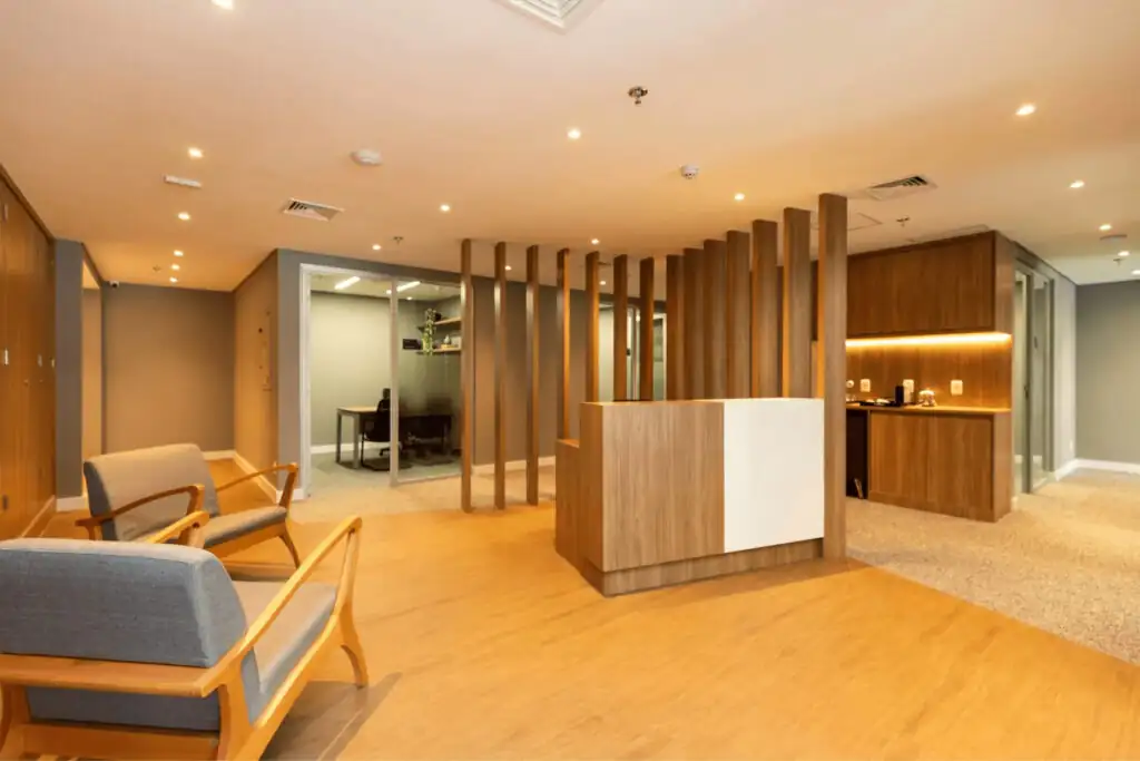 Recepção do escritório Villemor Amaral com balcão em madeira, divisórias verticais decorativas, poltronas aconchegantes e iluminação quente, destacando projeto de arquitetura e reforma corporativa de alto padrão.