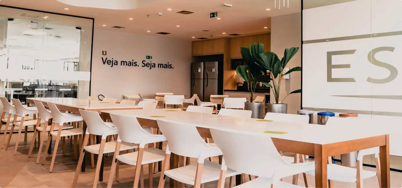 Área de convivência do escritório da Essilor no Rio de Janeiro, com mesas brancas, cadeiras de madeira, cozinha integrada e ambiente corporativo iluminado, mostrando soluções de arquitetura para escritórios modernos.