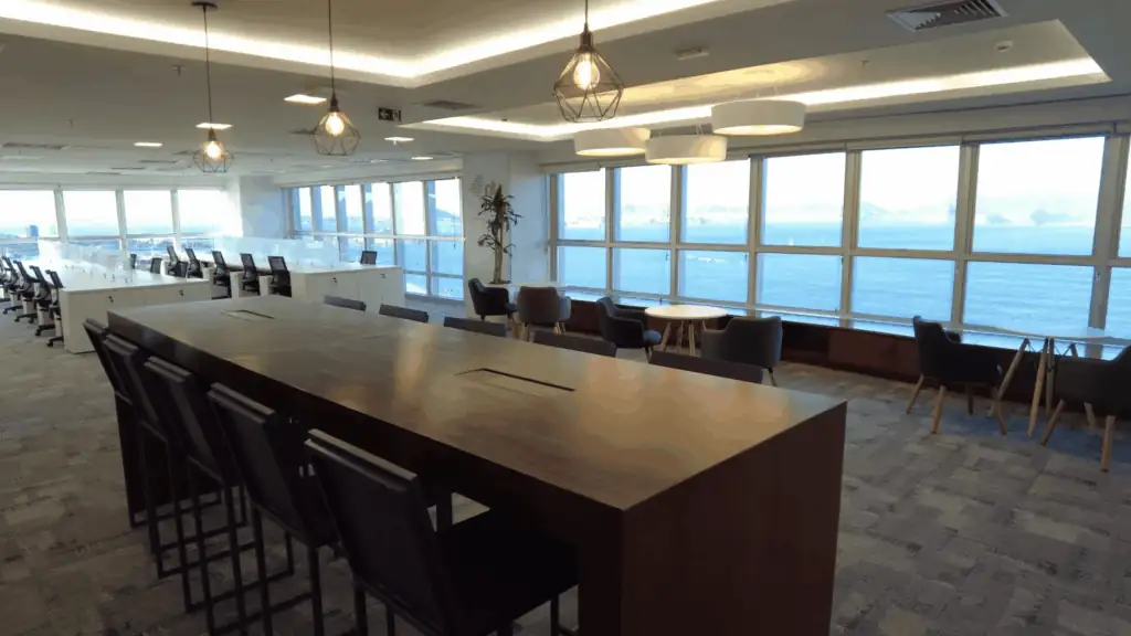 Área de convivência do escritório da NTS com grandes janelas voltadas para o mar, mesas de apoio, bancada alta com cadeiras e iluminação decorativa, representando um espaço planejado por escritório de arquitetura. A ambientação faz parte de um projeto de arquitetura pensado para bem-estar e integração, resultado de uma obra corporativa completa e reforma de escritório que valoriza vista, conforto e funcionalidade.