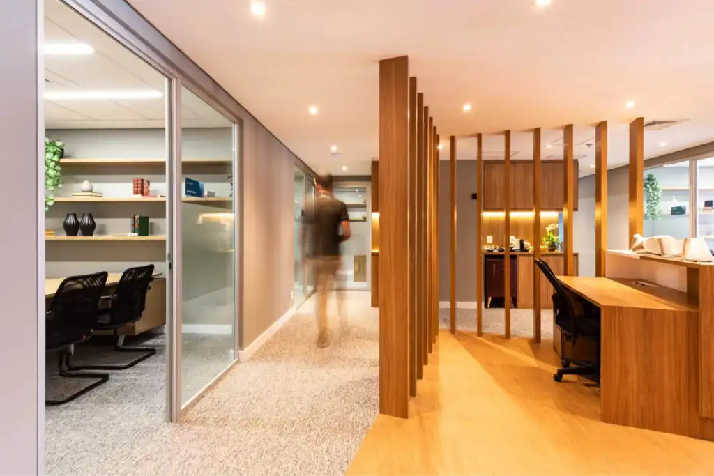 Corredor do escritório Villemor Amaral com divisórias verticais em madeira, recepção integrada, salas envidraçadas e detalhes de iluminação aconchegante, destacando projeto de arquitetura e reforma corporativa de alto padrão.