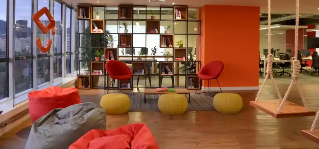 Espaço de descompressão do escritório da Passei Direto. Projeto de arquitetura com puffs coloridos, balanços, poltronas vermelhas e estante decorativa, em reforma corporativa moderna e descontraída com ampla vista para a cidade.