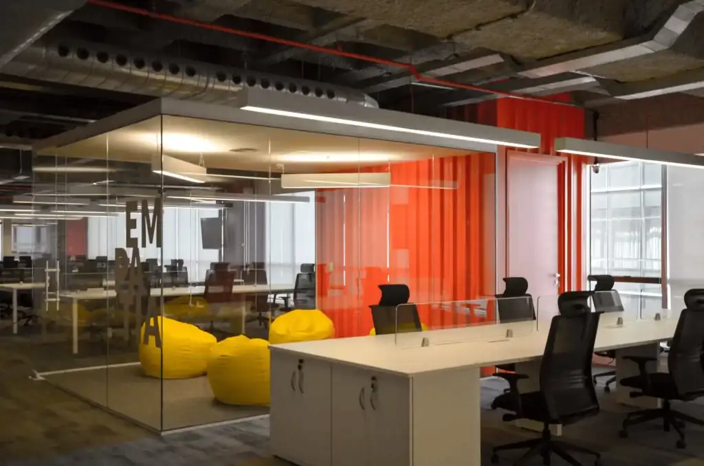 Área colaborativa da Passei Direto com sala de vidro, puffs amarelos e estação de trabalho moderna, desenvolvida por escritório de arquitetura. Projeto de arquitetura focado em obra corporativa e reforma de escritório, com elementos industriais e divisórias coloridas.