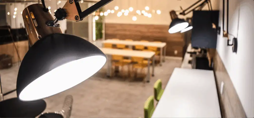 Área de descompressão do escritório da Pebmed com luminárias articuladas em destaque e mesas ao fundo, mostrando um ambiente moderno criado através de projeto de arquitetura e obra corporativa, com foco em funcionalidade e bem-estar no espaço de trabalho.