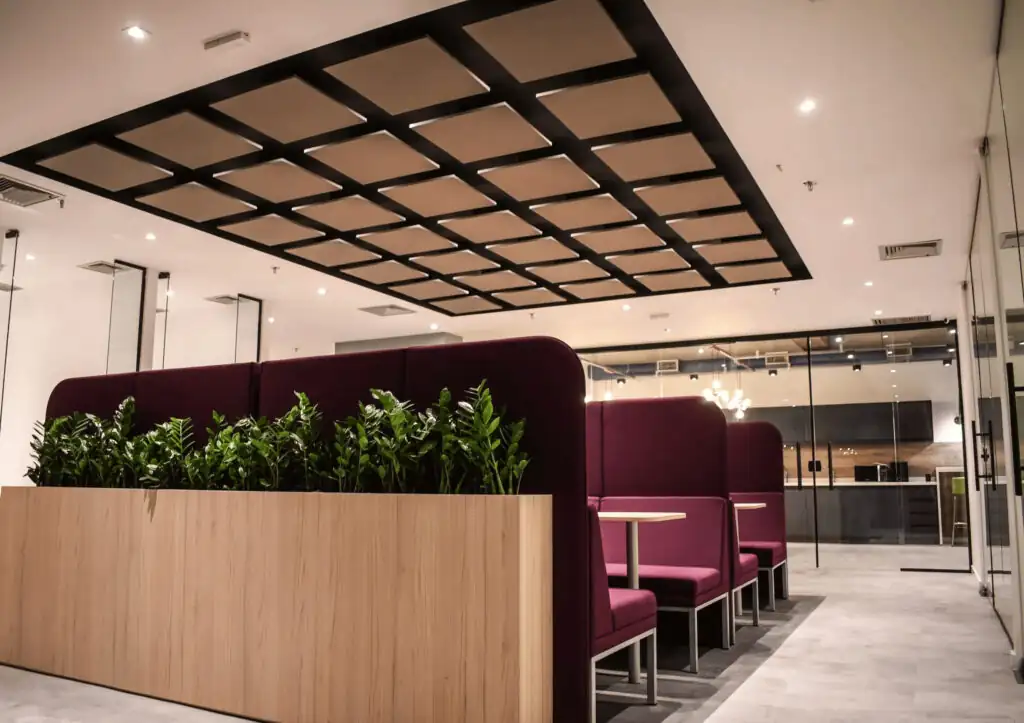 Espaço colaborativo do escritório da Pebmed com cabines estofadas na cor vinho e divisórias com vegetação, integrando elementos modernos de projeto de arquitetura e reforma de escritório corporativo em um ambiente sofisticado.