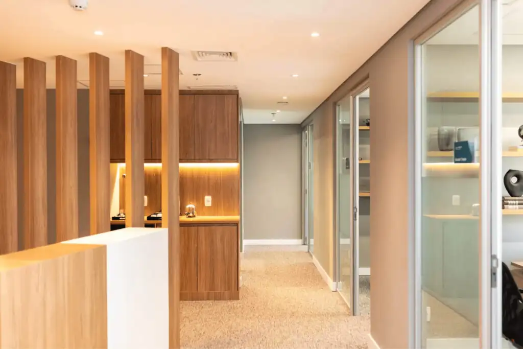 Corredor do escritório Villemor Amaral com divisórias de madeira, salas envidraçadas e marcenaria iluminada, destacando projeto de arquitetura sofisticado e reforma corporativa de alto padrão.