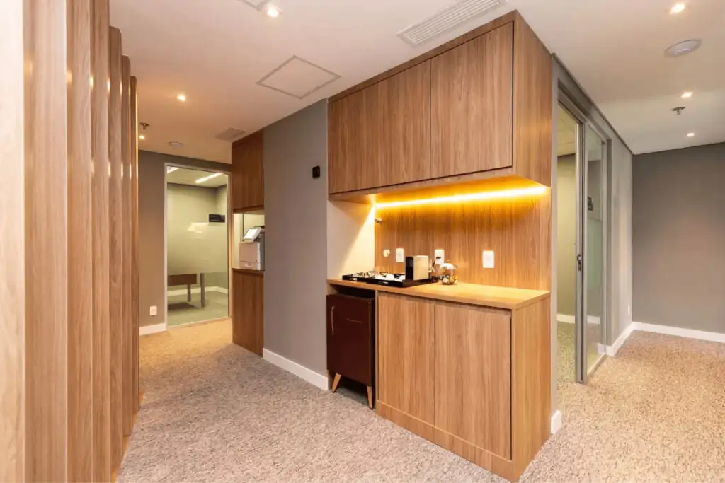 Área de copa do escritório Villemor Amaral com marcenaria em madeira, iluminação embutida e corredor integrado a salas envidraçadas, destacando obra corporativa e projeto de arquitetura elegante e funcional.