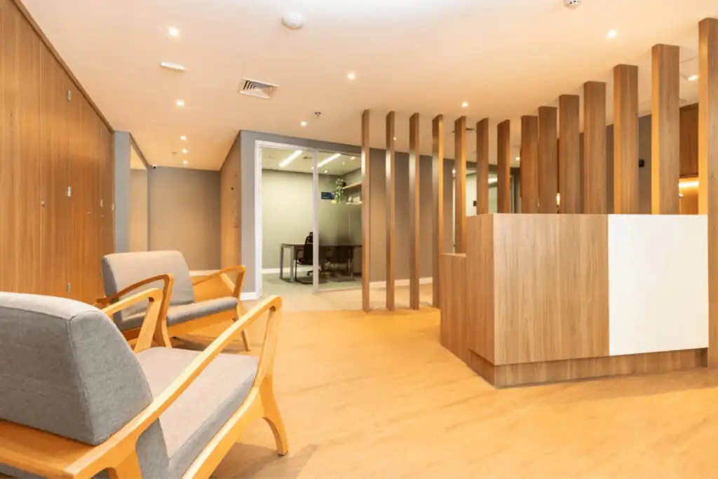 Recepção do escritório Villemor Amaral com cadeiras de madeira e tecido cinza, elementos verticais em madeira e sala envidraçada ao fundo, destacando projeto de arquitetura corporativa e reforma de escritório de alto padrão.