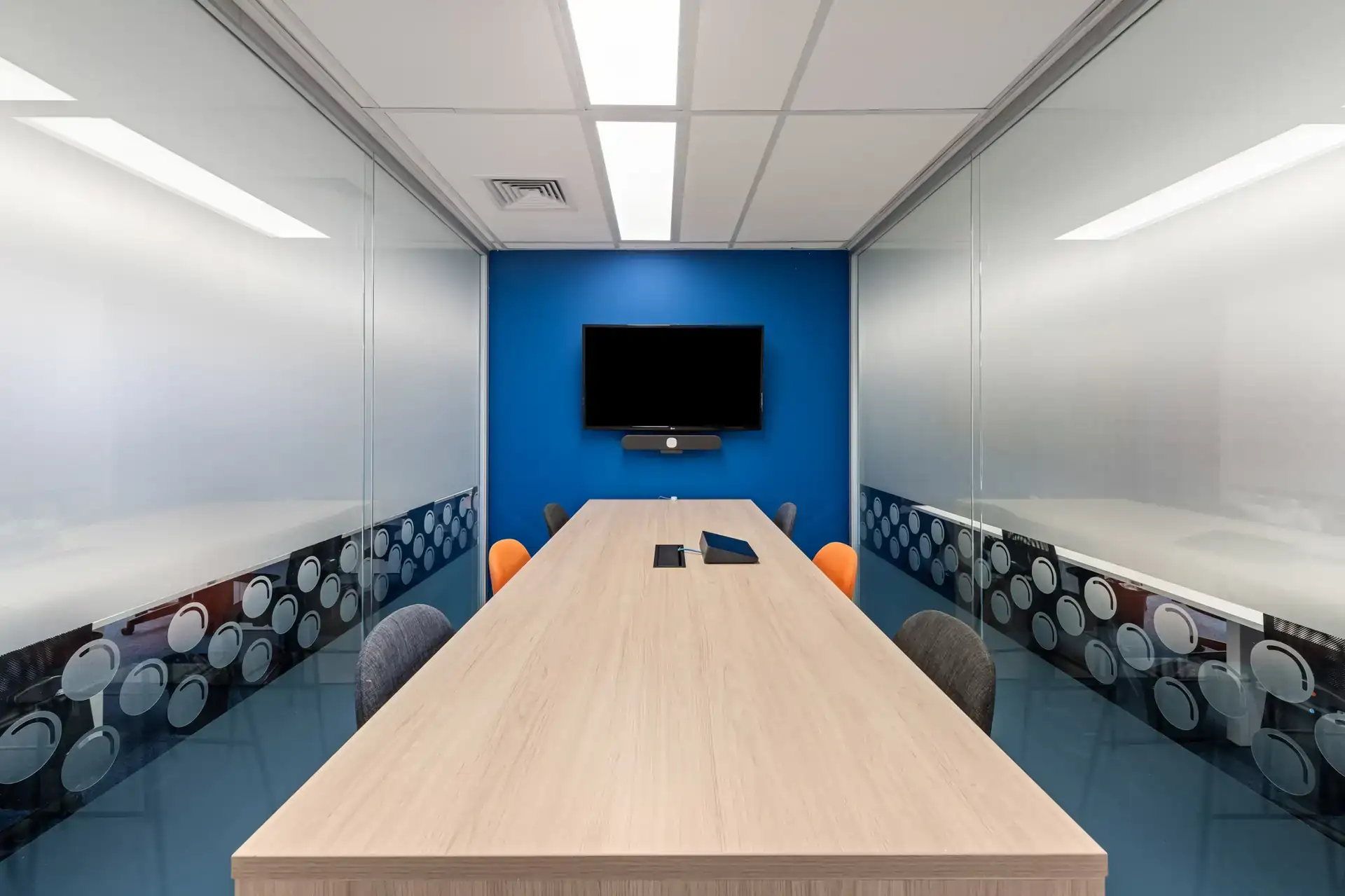Sala de reunião compacta desenvolvida em projeto de arquitetura corporativa, com mesa de madeira, divisórias de vidro e parede azul para videoconferência.