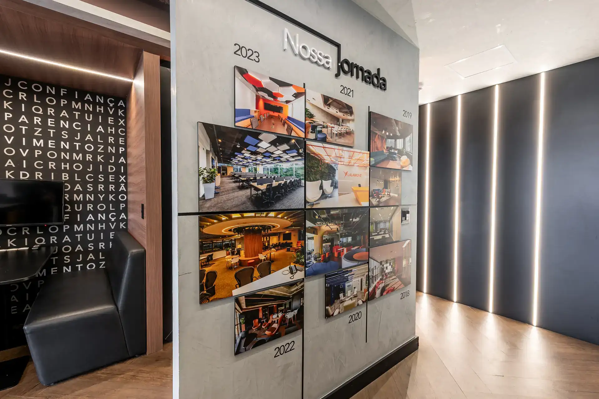 Painel “Nossa Jornada” exibindo projetos de arquitetura entregues ao longo dos anos, integrado ao corredor com iluminação linear moderna em ambiente de obra corporativa finalizada.