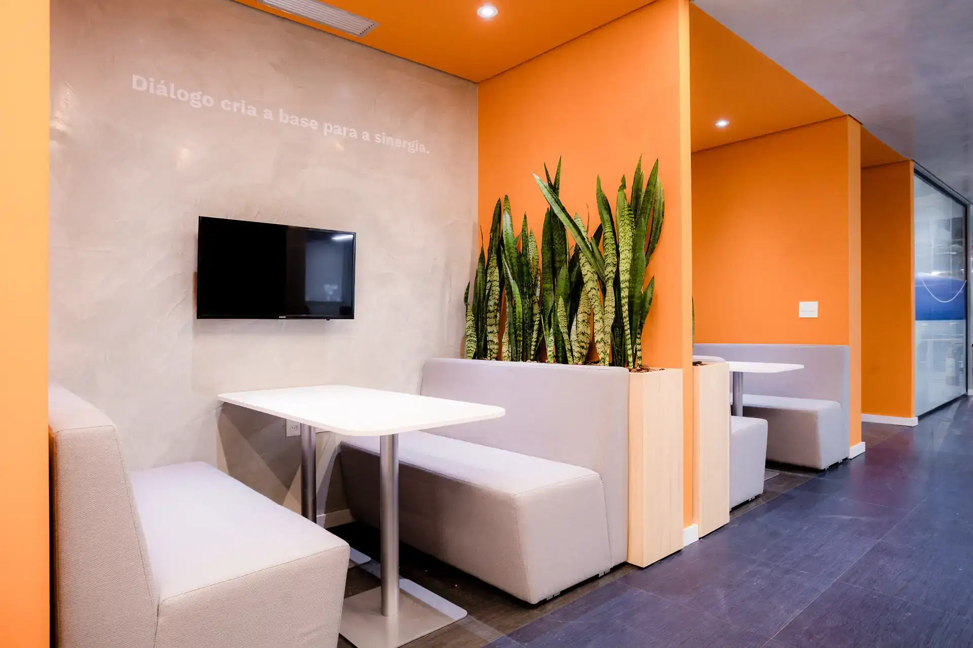 Booths colaborativos no escritório da EDF Macaé com estofados cinza, paredes laranja e vegetação. Espaço criado em reforma corporativa para diálogos rápidos e reuniões informais.