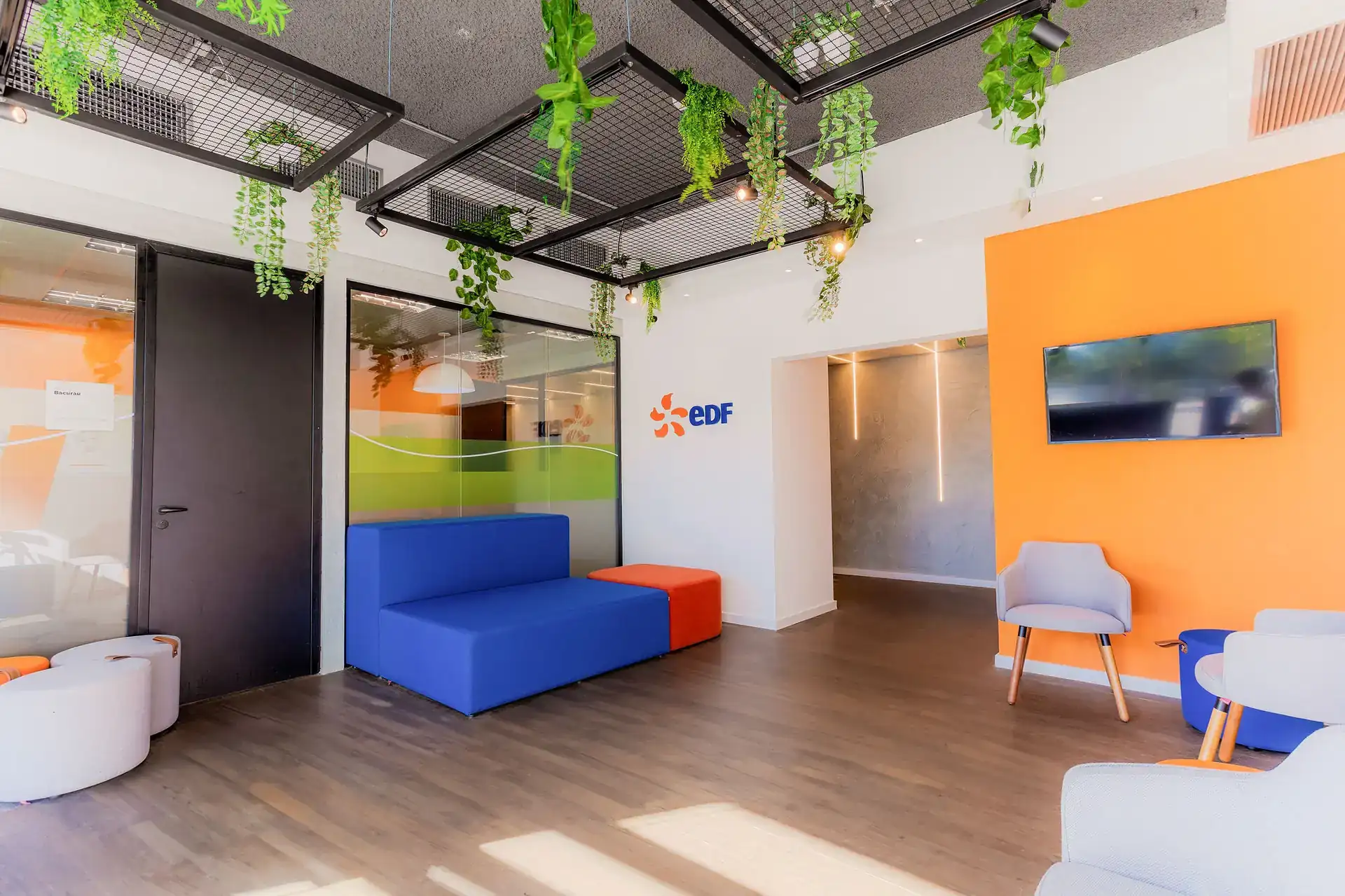 Recepção da EDF Macaé com teto metálico vegetado, mobiliário azul e laranja, e logo EDF na parede. Projeto de arquitetura que destaca a identidade visual da empresa.