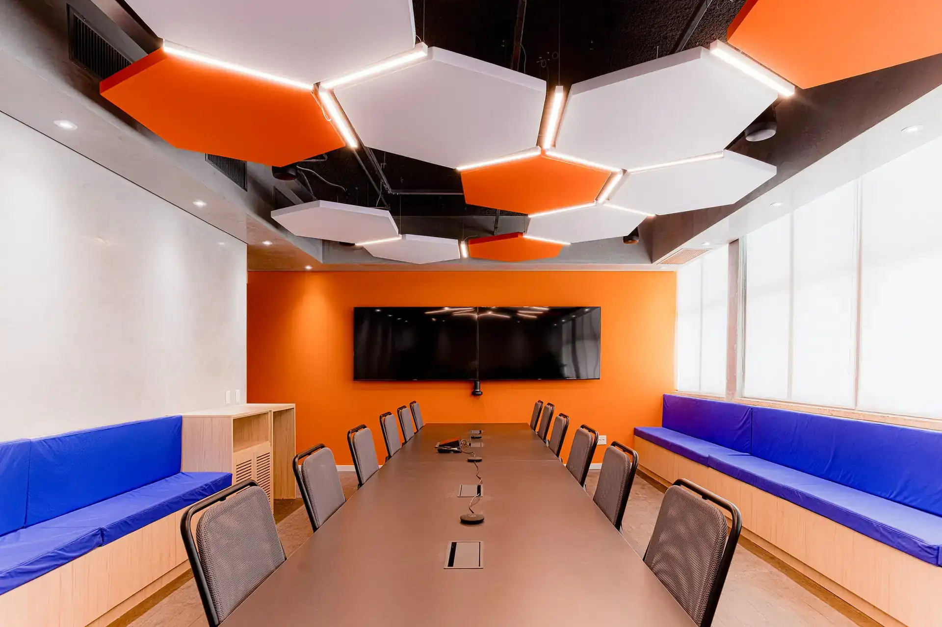 Sala de reunião no escritório corporativo da EDF Macaé com teto geométrico iluminado, mesa central escura e bancos laterais azuis. Projeto de arquitetura corporativa com foco em funcionalidade e estética vibrante.