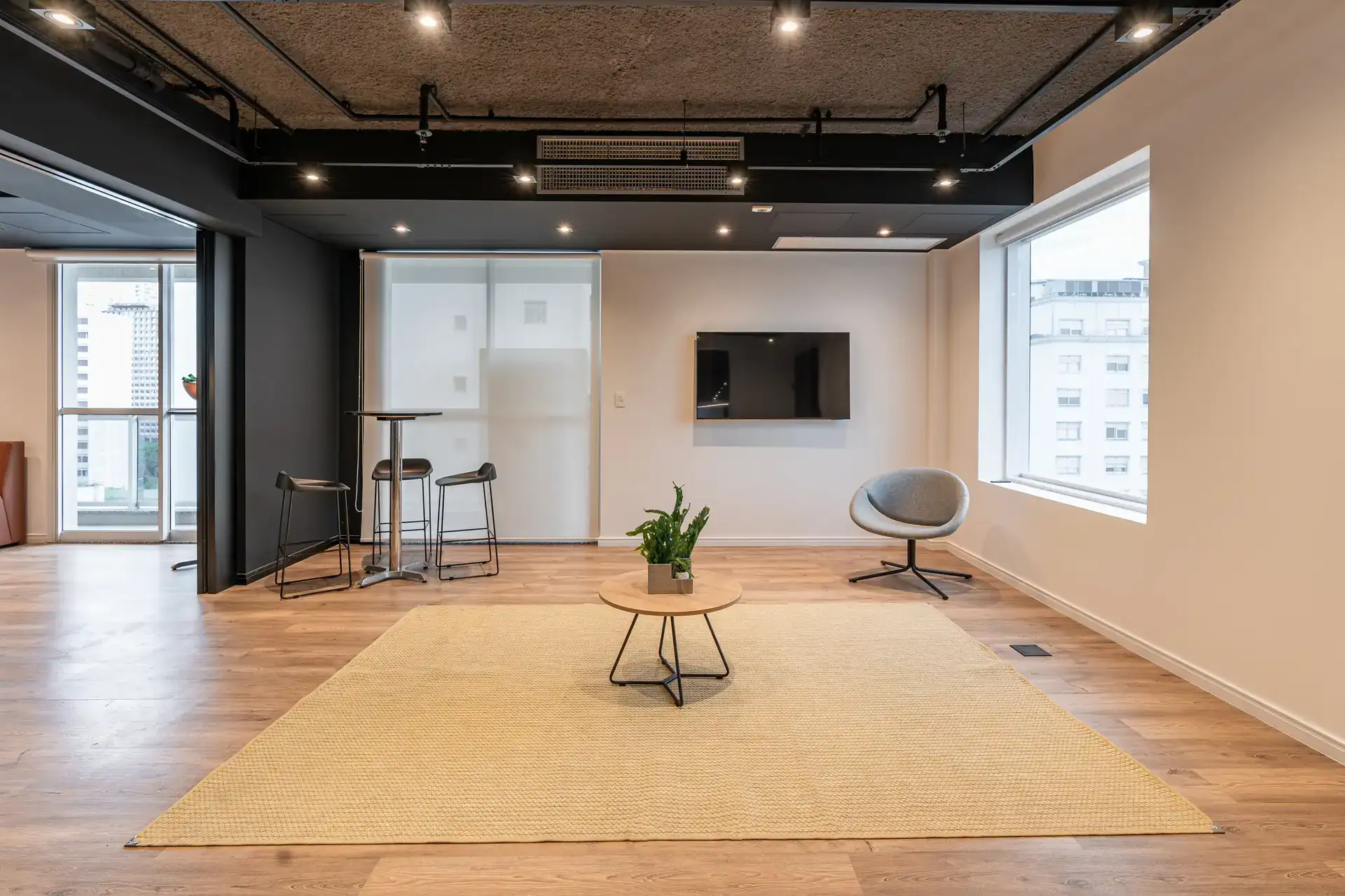 Área multiuso com tapete claro, mesa central e janelas amplas, projetada para eventos e convivência dentro do escritório corporativo.