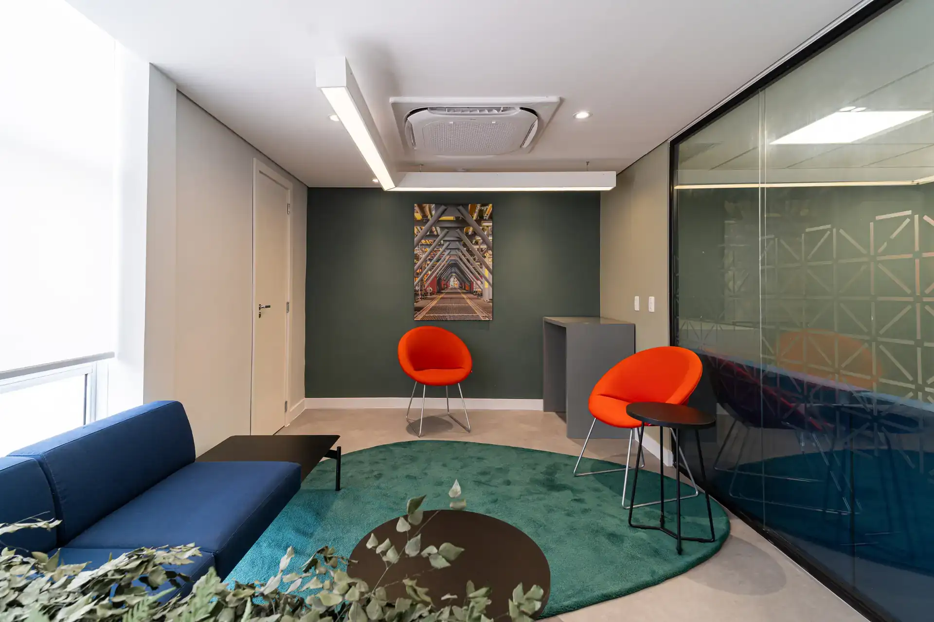 Lounge corporativo com poltronas laranjas e azul, tapete verde e iluminação suave, integrado ao projeto de arquitetura do escritório da NSG.