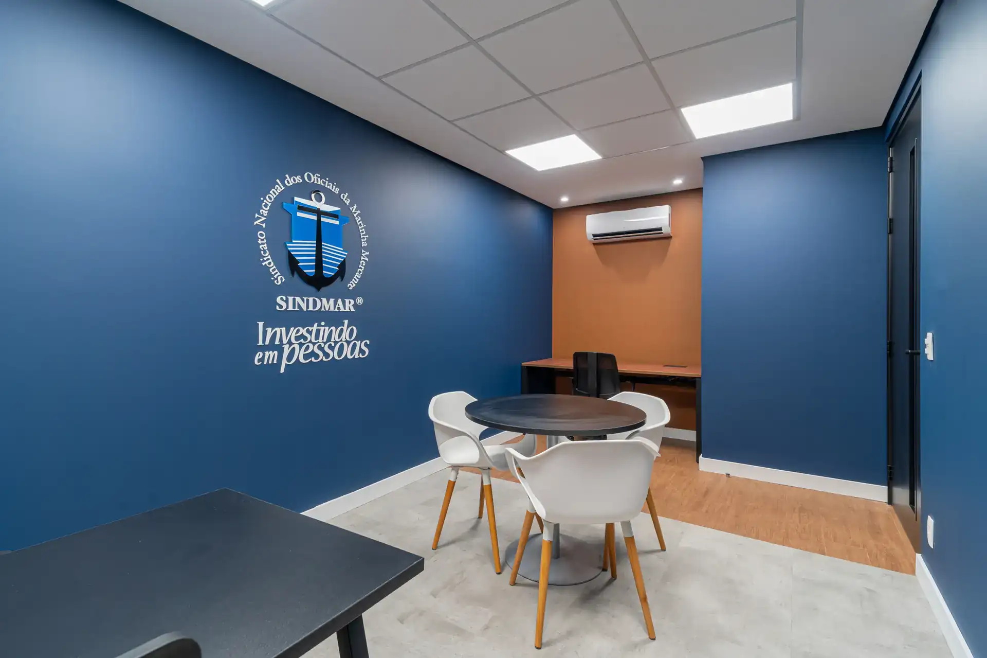 Sala de reuniões compacta com mesa redonda e logo no escritório do SINDMAR em parede azul, entregue em projeto de arquitetura e obra corporativa.