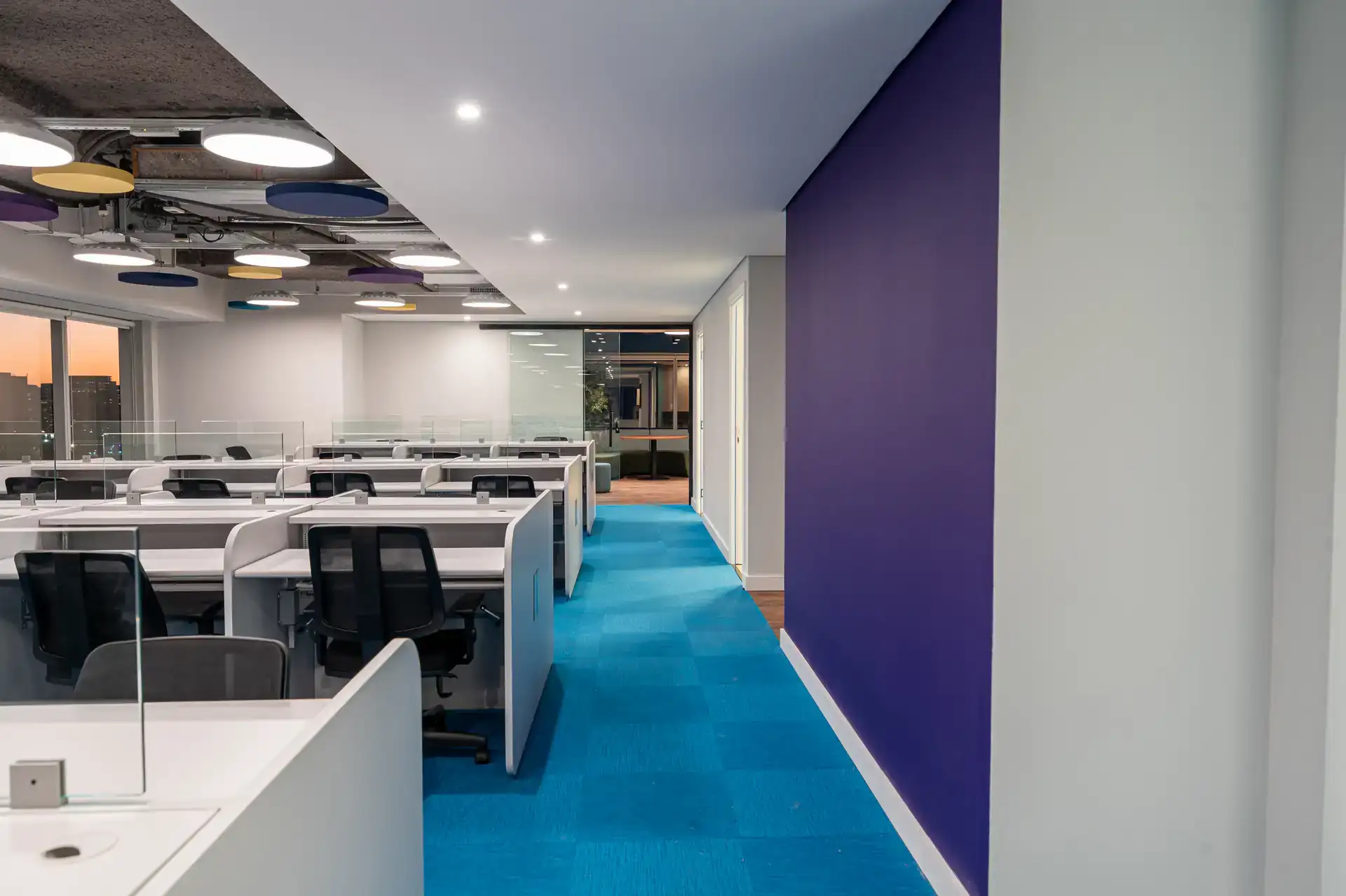 Open space do escritório da Inove com corredor azul, parte do projeto de arquitetura e obra corporativa entregue, com estações de trabalho, divisórias em vidro e teto industrial com painéis circulares coloridos.