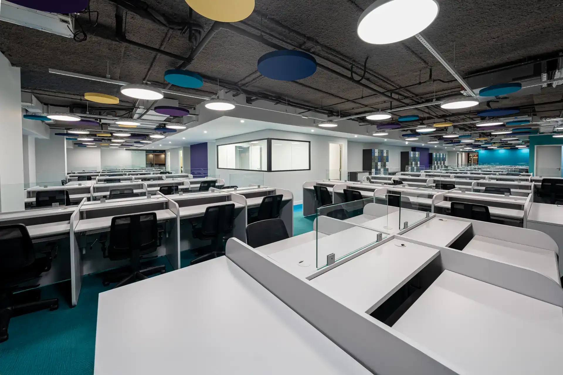 Open space do escritório da Inove após reforma corporativa entregue, com estações de trabalho amplas, divisórias em vidro, teto aparente e elementos coloridos.