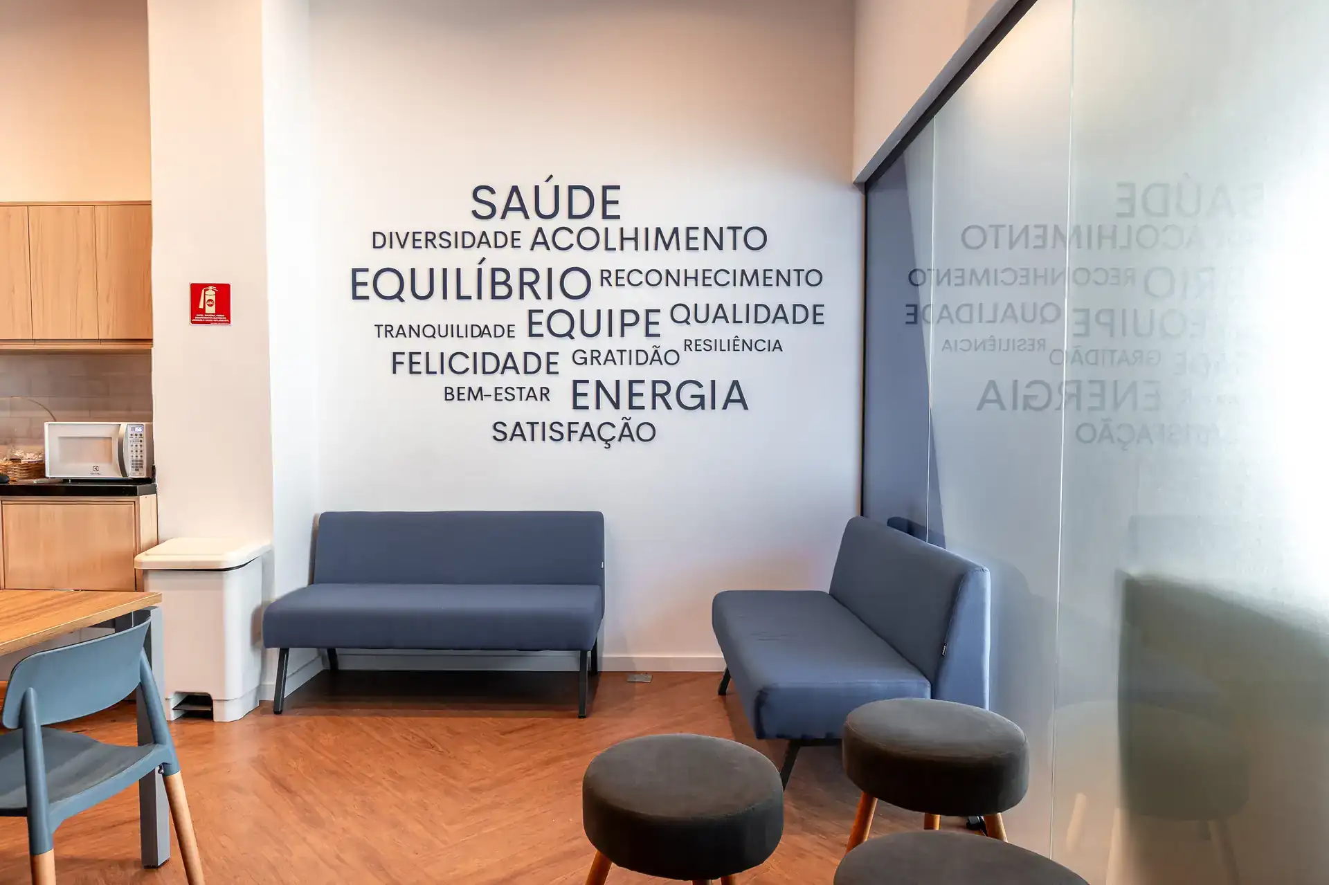 Área de descompressão com sofás azuis, bancos e parede com palavras sobre saúde e equilíbrio, resultado de reforma de escritório voltada ao bem-estar da equipe.