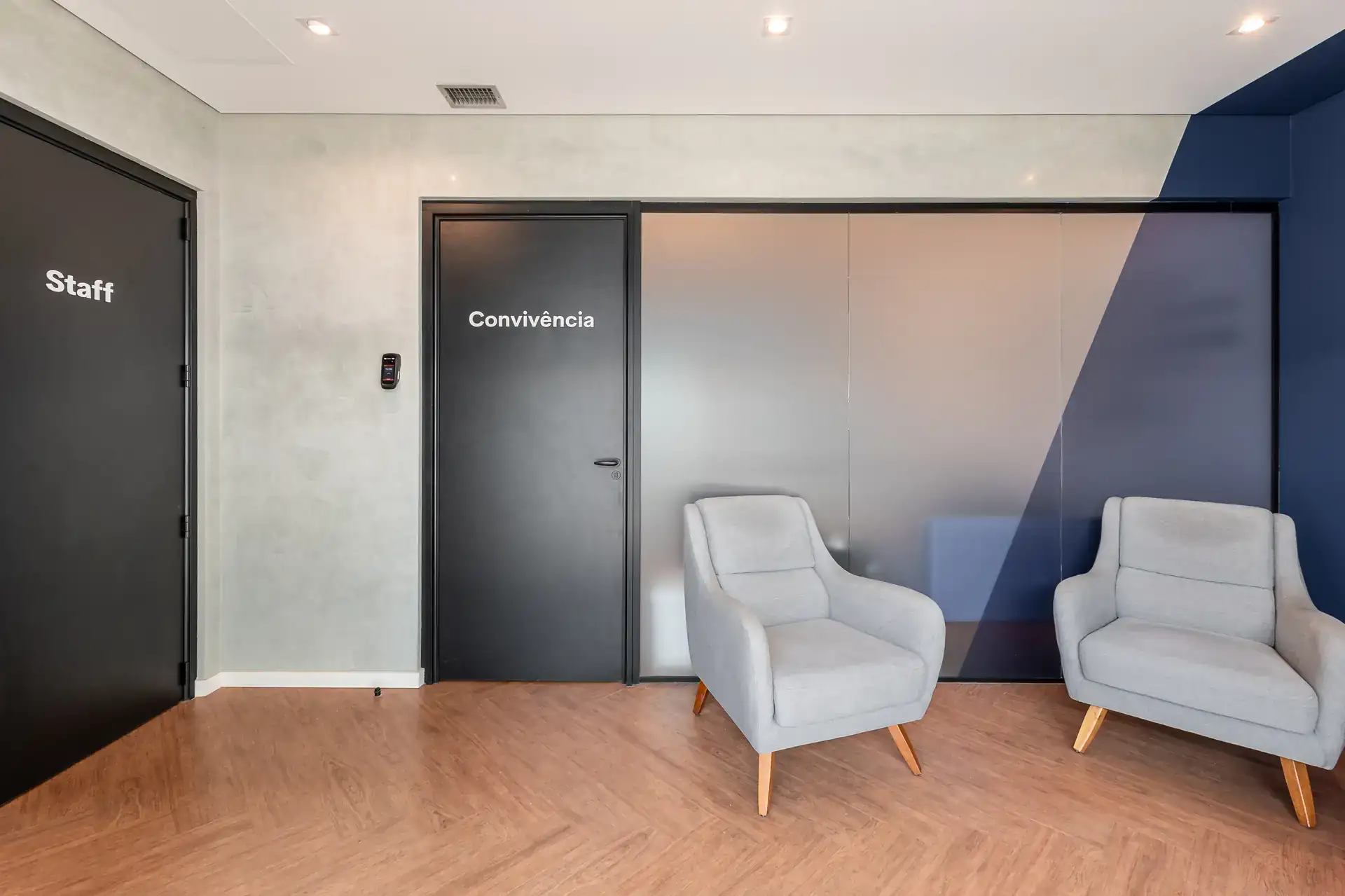 Lobby de convivência com poltronas cinza, portas sinalizadas e piso amadeirado, integrando áreas de staff e convivência em projeto de arquitetura corporativa para o escritório da LAM Medical.