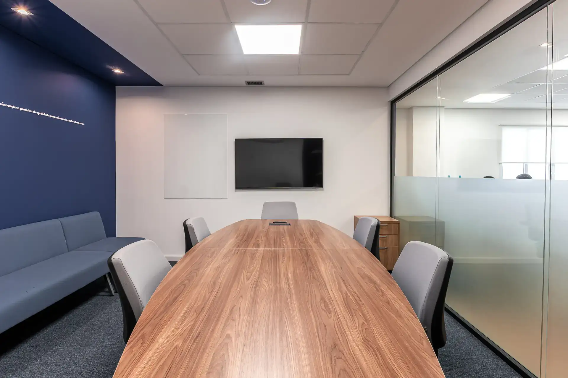 Sala de reunião com mesa oval em madeira, cadeiras cinza e TV frontal, criada em obra corporativa que valoriza conforto acústico e privacidade.