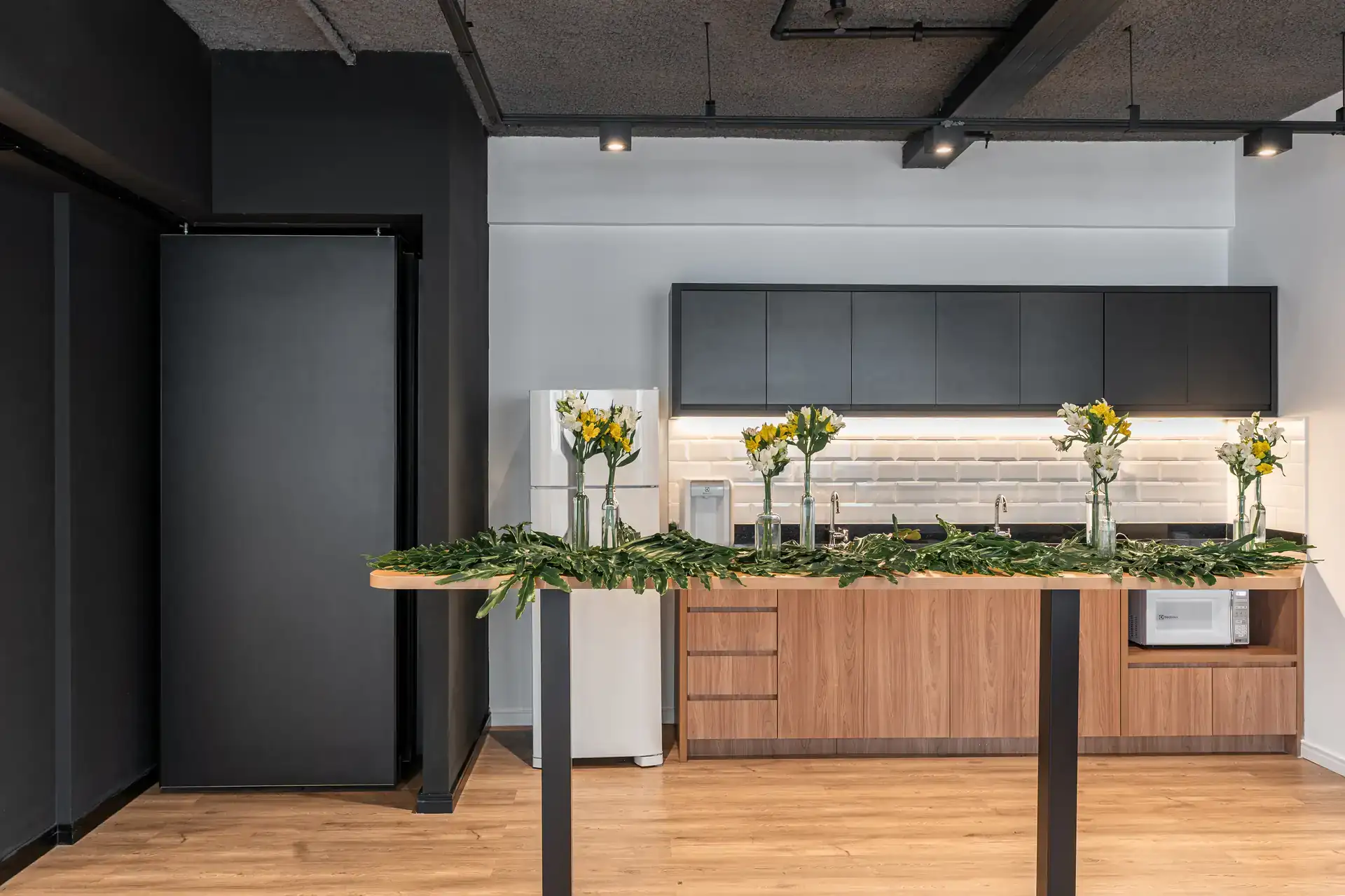 Copa corporativa com bancada em madeira, armários pretos e decoração com flores, parte da reforma do escritório da MHM.