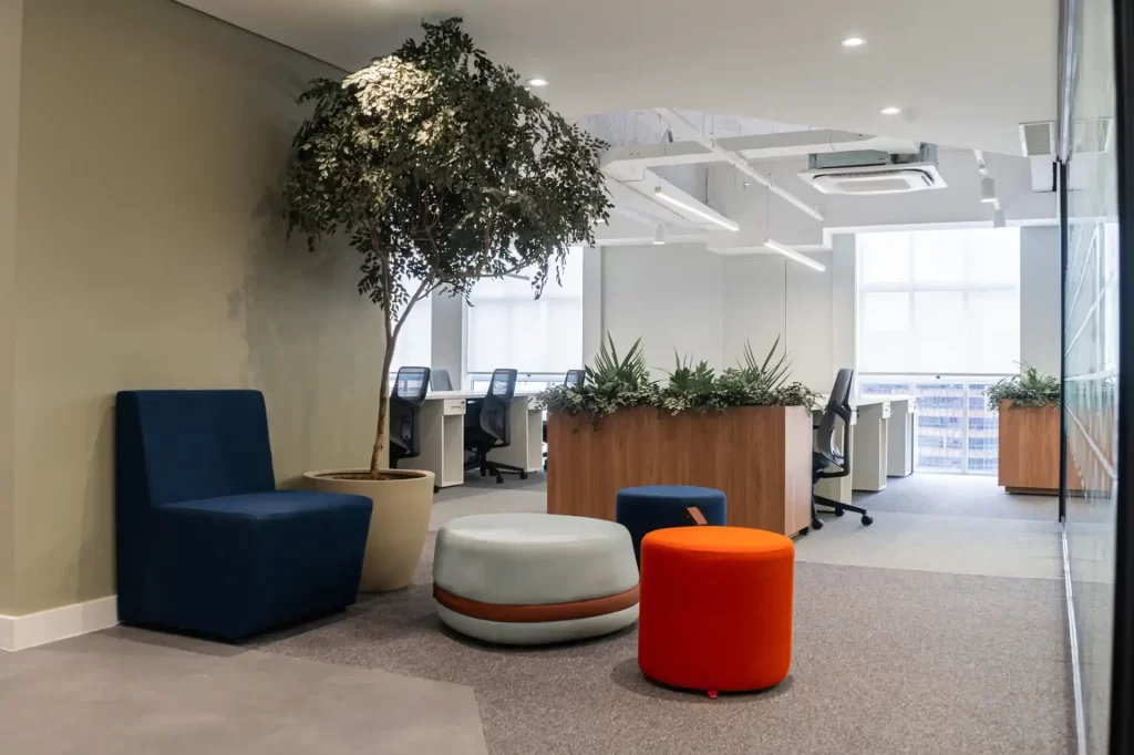 Espaço de descompressão com mobiliário colorido, vegetação e integração ao open space, evidenciando reforma de escritório que prioriza bem-estar e conforto dos colaboradores com arquitetura corporativa.