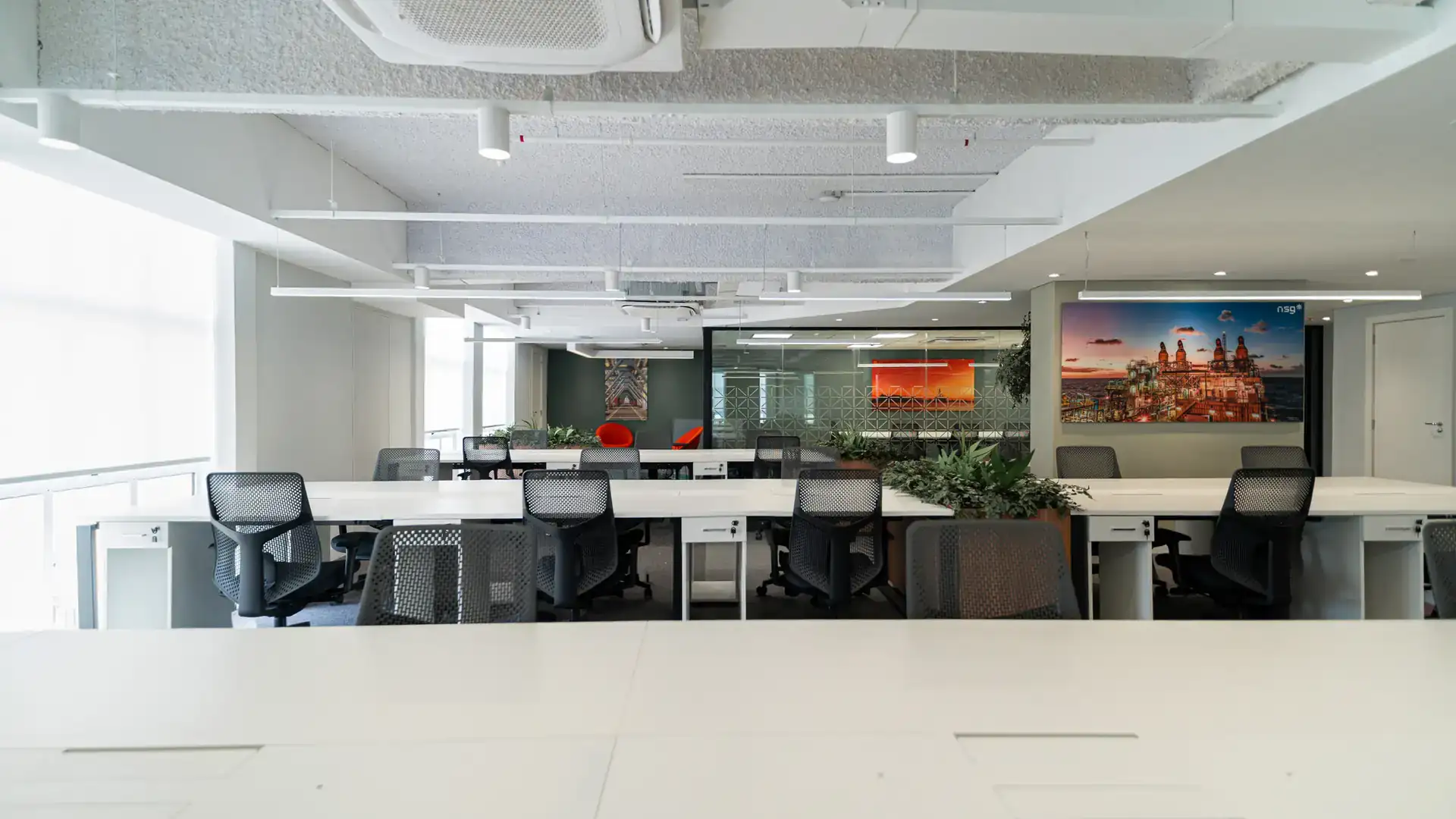 Área ampla com estações de trabalho alinhadas, iluminação moderna e integração entre escritório aberto e sala envidraçada, destacando soluções de projeto de arquitetura corporativa.