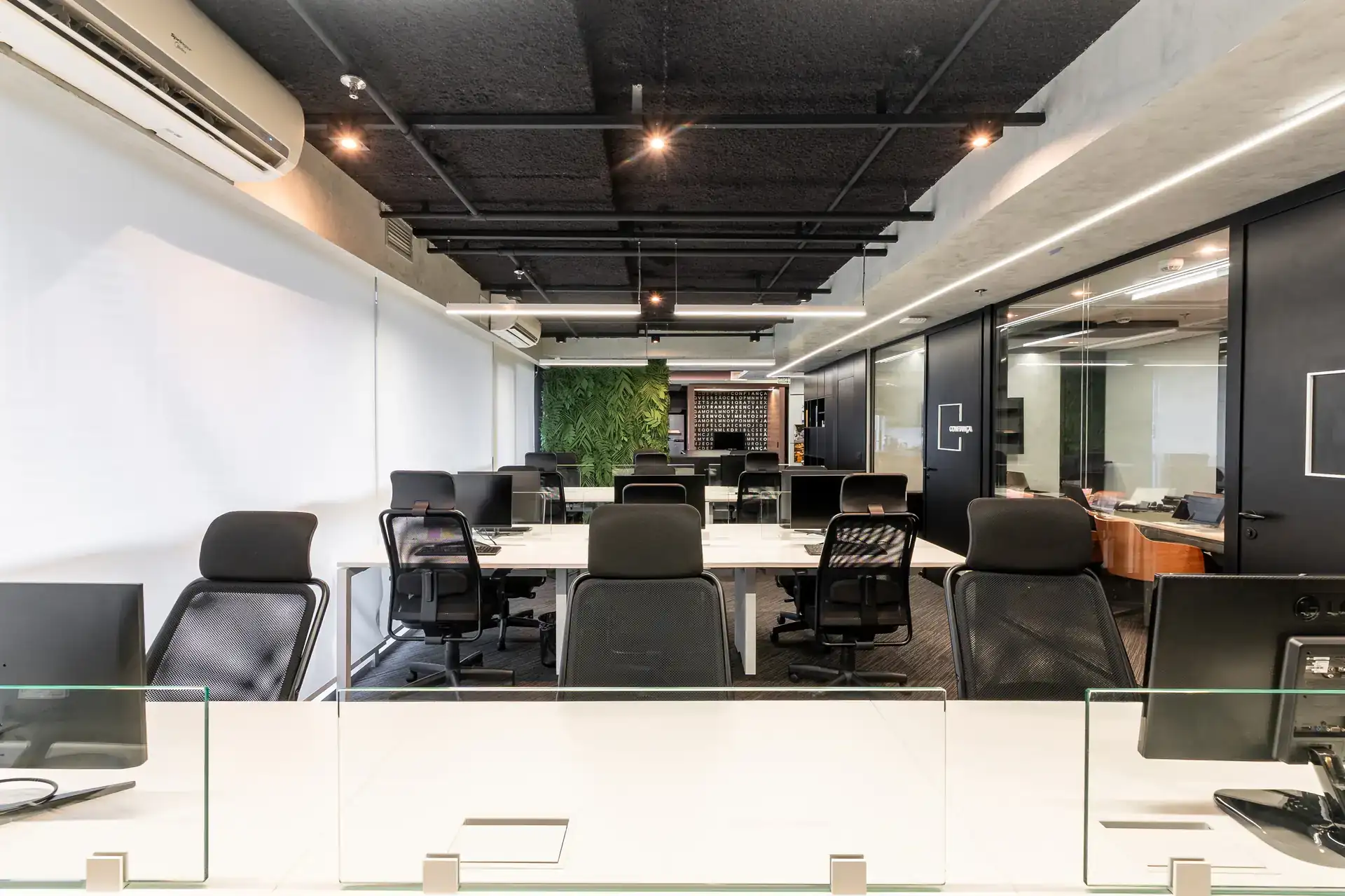 Estações de trabalho integradas em obra corporativa da Custódio, com design industrial, iluminação embutida e painel verde ao fundo. Reforma de escritório executada com padrão premium de arquitetura.