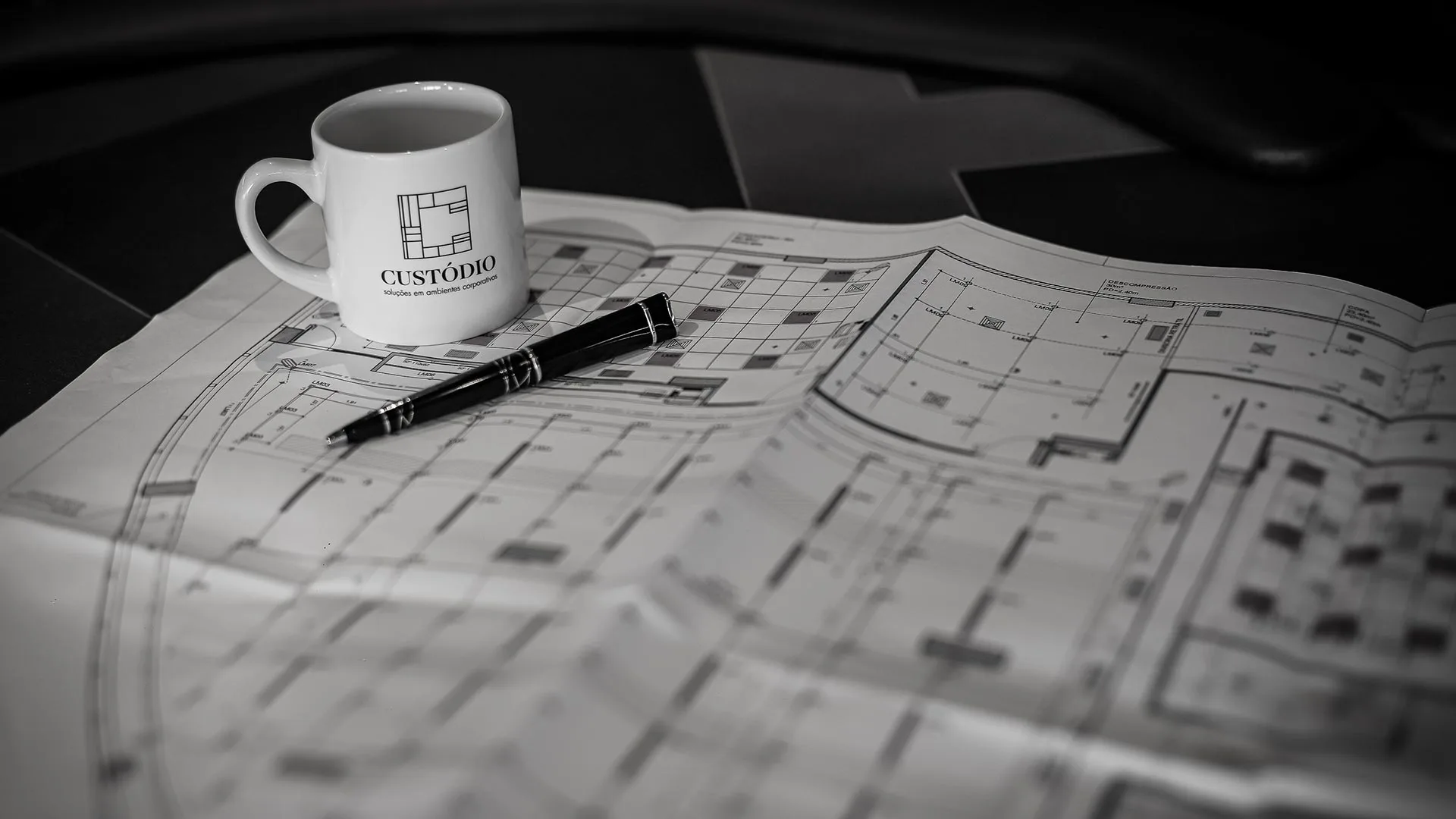 Foto institucional da Custódio mostrando planta arquitetônica e caneca da marca sobre mesa de projeto. Representa o processo de criação e detalhamento técnico em projetos e obras corporativas de alto padrão.