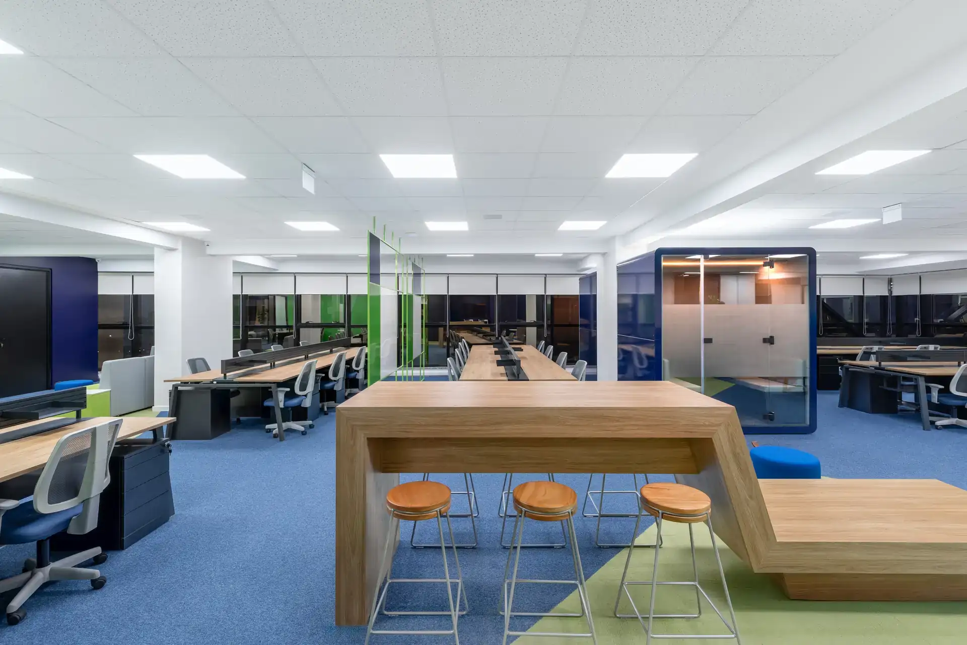 Bancada colaborativa em madeira integrada ao open space, com estações de trabalho e phonebooth ao fundo, destacando o projeto de arquitetura e a reforma corporativa da Transit BR.